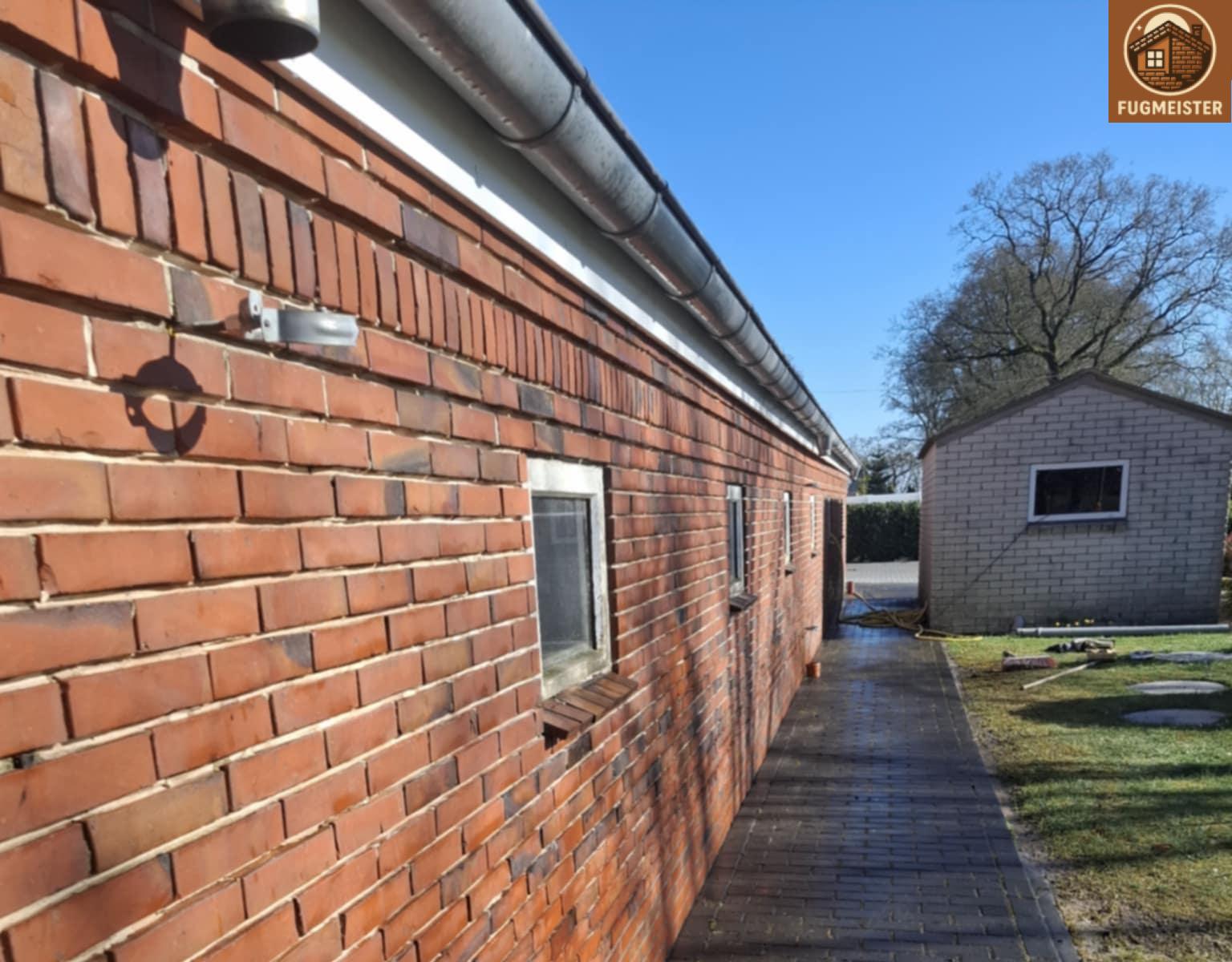 Lange Seitenwand aus roten Klinkern mit kleinen Fenstern, Dachrinne und Hofweg im Sonnenlicht.