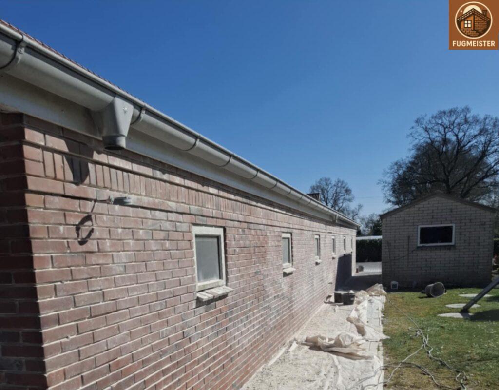Lange Hausfassade aus Klinker mit Dachrinne und Hofseite; gereinigte Wandabschnitte und Gartenhaus im Hintergrund.