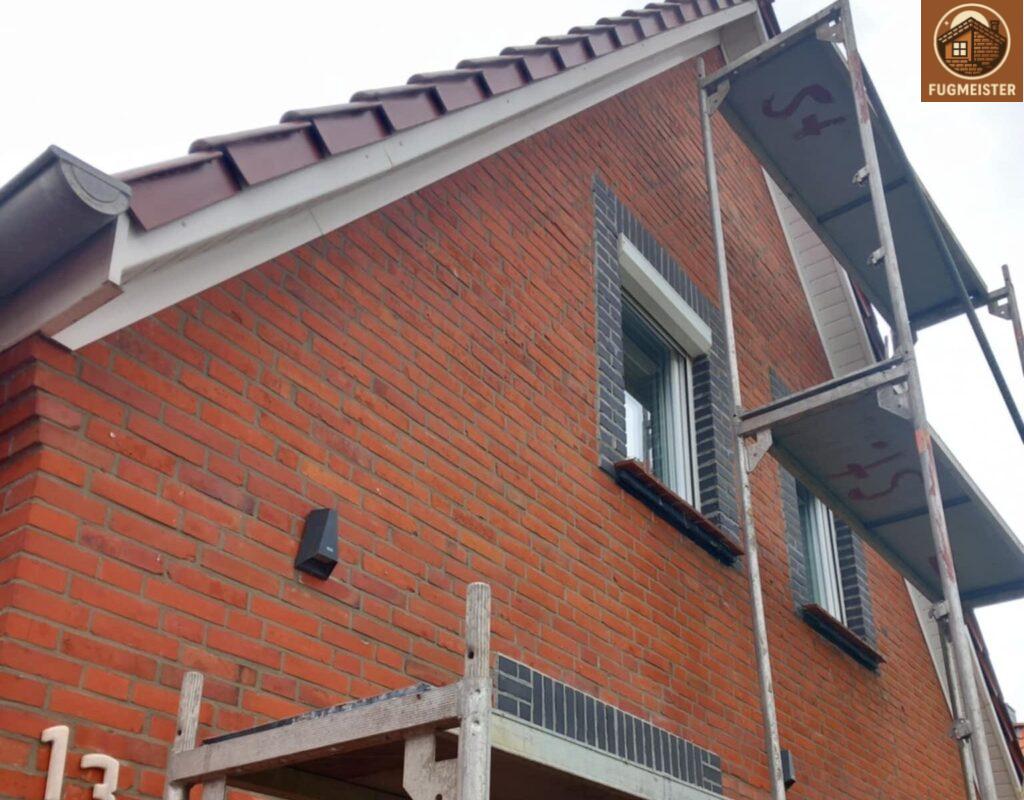 Seitenansicht eines Klinkerhauses mit roten Ziegeln, dunklen Fensterlaibungen und Arbeitsgerüst für Fassadenarbeiten.